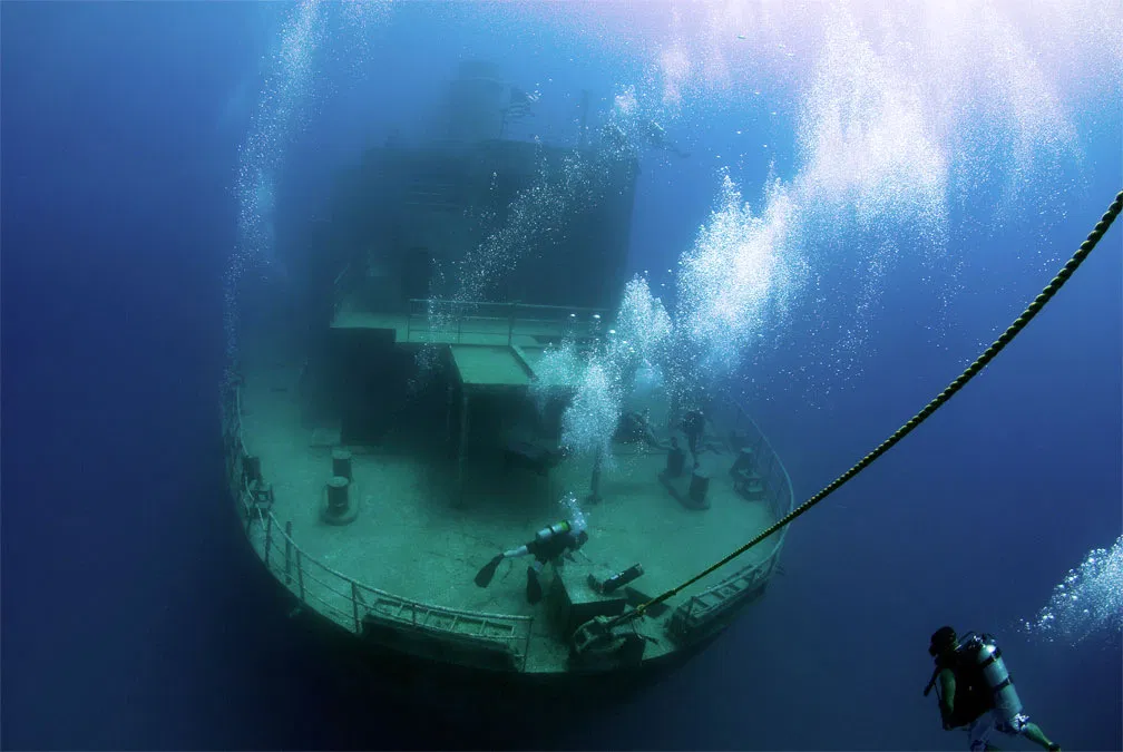 Divers descending on a large shipwreck in blue water, bubble columns rising from the deck.